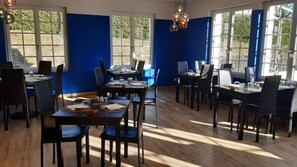 A dining area with multiple tables and chairs, large windows with a view of greenery, and a wooden floor. - Logis Hotel le 1050 (Saint-Jeure-d'Andaure)