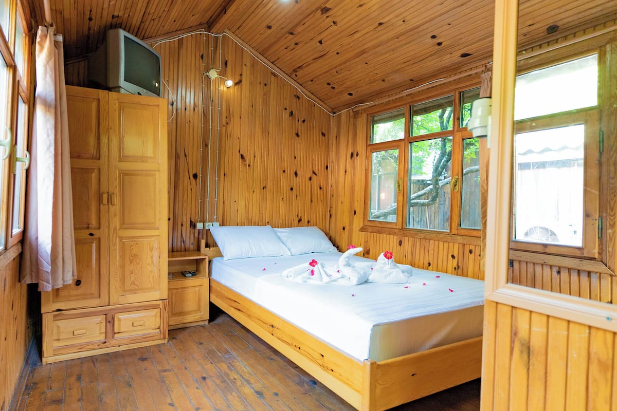 A wooden cabin room with a bed, a TV on a cabinet, and a window with a view of trees.