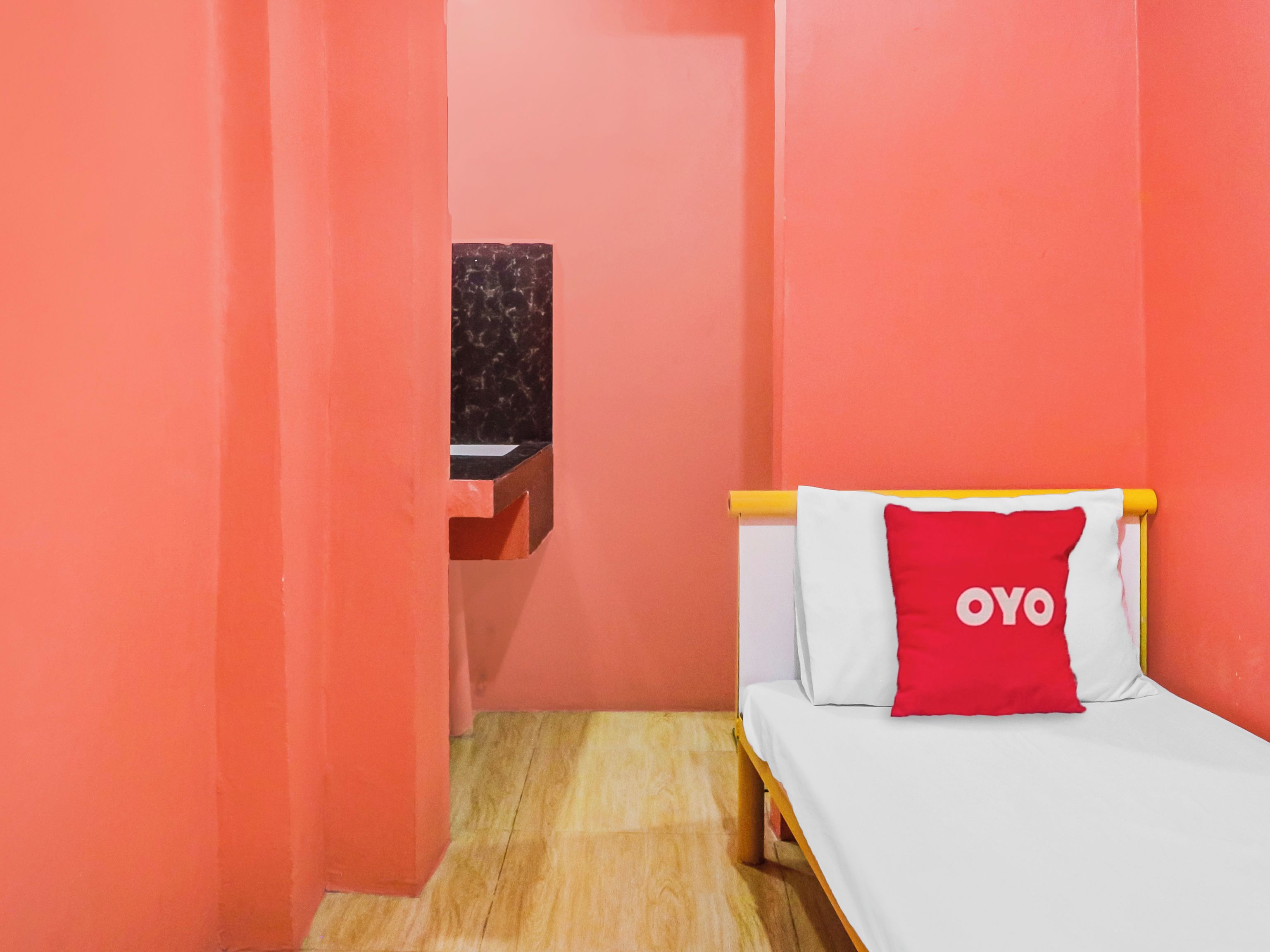 Ein kleines Zimmer mit einem Bett, einem roten Kopfkissen mit der Aufschrift 'OYO', einem hölzernen Kopfteil und einem kleinen Tisch mit schwarzer Marmorplatte.