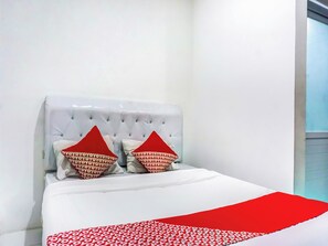 Uma cama com cabeceira estofada, duas almofadas vermelhas com padrão repetido e um edredom branco.