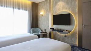 Habitación de hotel con dos camas, un televisor montado en la pared, una mesita con una cafetera, una silla y un amplio ventanal con cortinas.