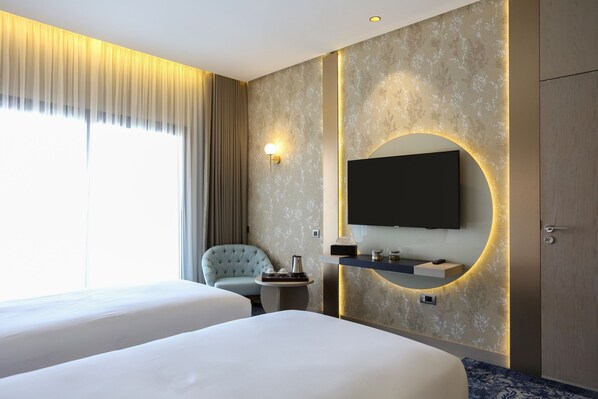 A hotel room with two beds, a TV mounted on the wall, a small table with a coffee pot, a chair, and a large window with curtains. - Oriental Palace (Taza)