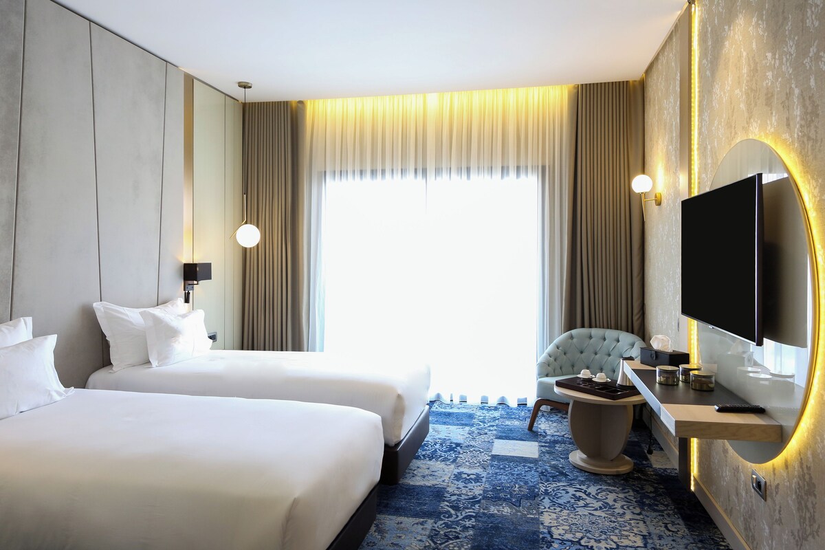 A hotel room with two beds, a desk with a TV, a chair, and a large window with curtains.