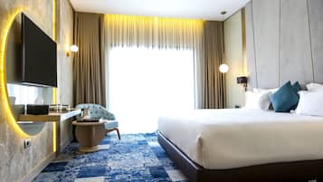 Ein Hotelzimmer mit einem großen Bett, einem an der Wand befestigten Fernseher, einem Schreibtisch mit Stuhl und einem großen Fenster mit Vorhängen.