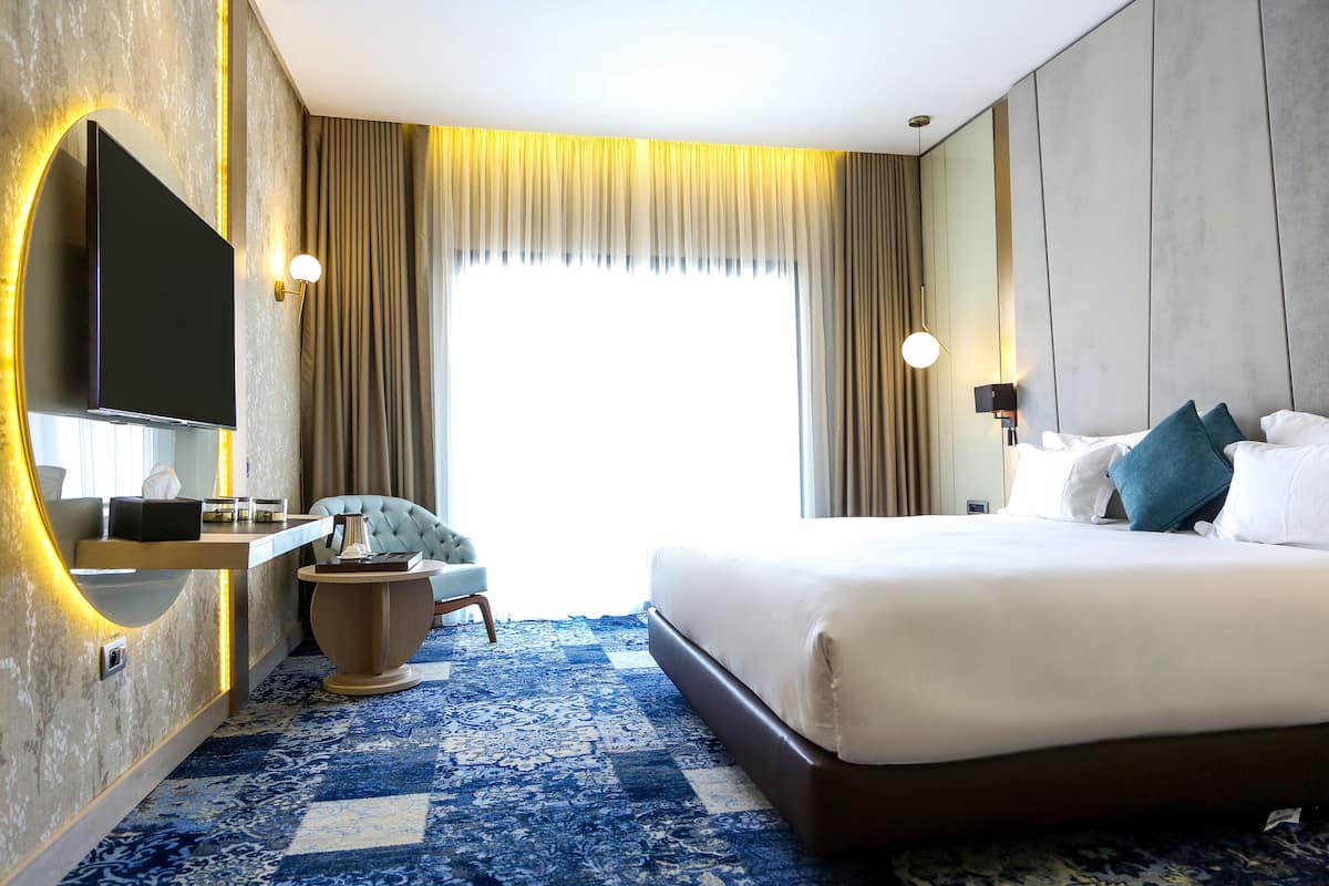 A hotel room with a large bed, a TV mounted on the wall, a desk with a chair, and a large window with curtains.