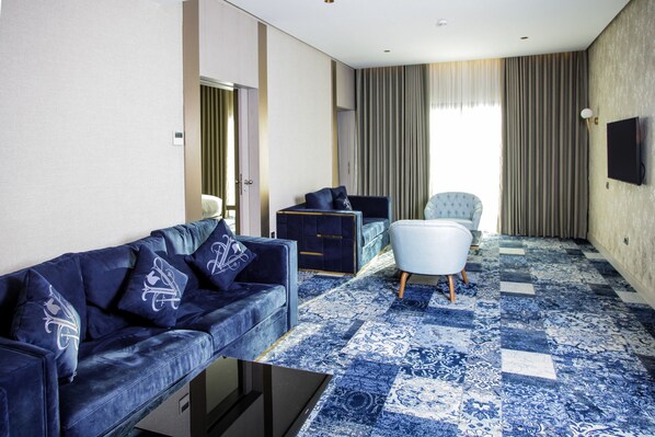 A modern hotel room with a blue patterned carpet, a dark blue sofa with decorative pillows, a white armchair, a small table, and a television mounted on the wall.