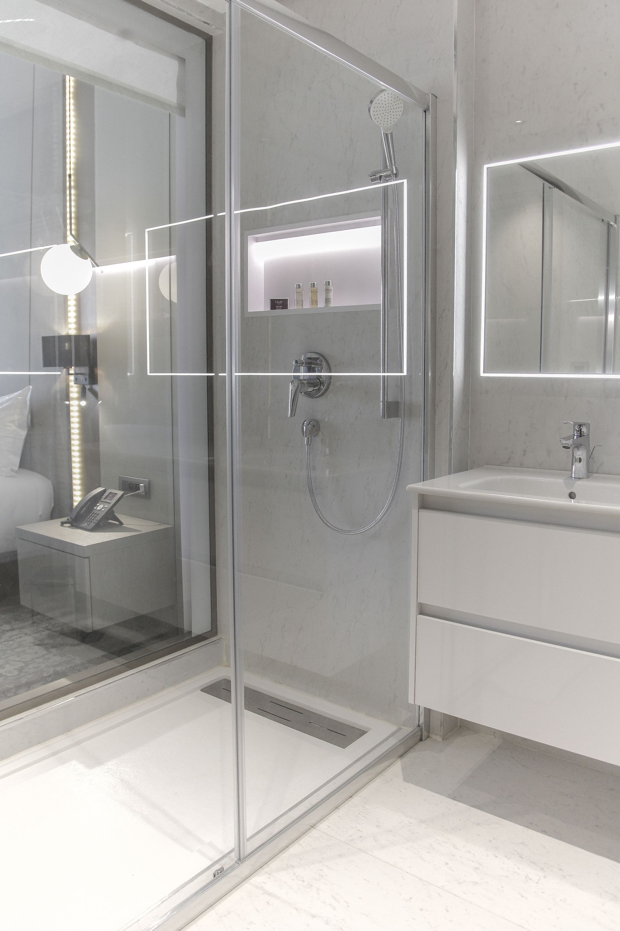 A modern bathroom with a glass-enclosed shower, a vanity with a mirror, and a bedside table with a lamp.