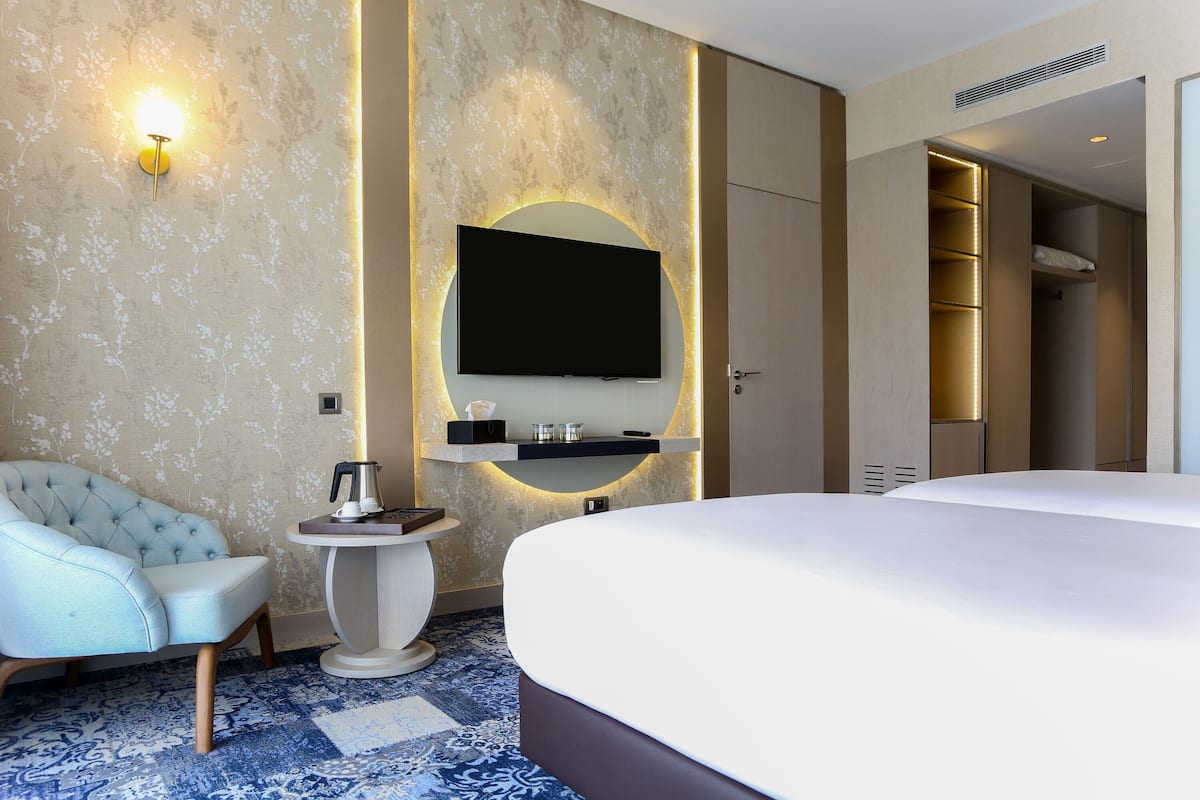 A hotel room with a bed, a seating area with a chair, a small table with a kettle, a flat-screen TV mounted on the wall, and a built-in shelving unit.