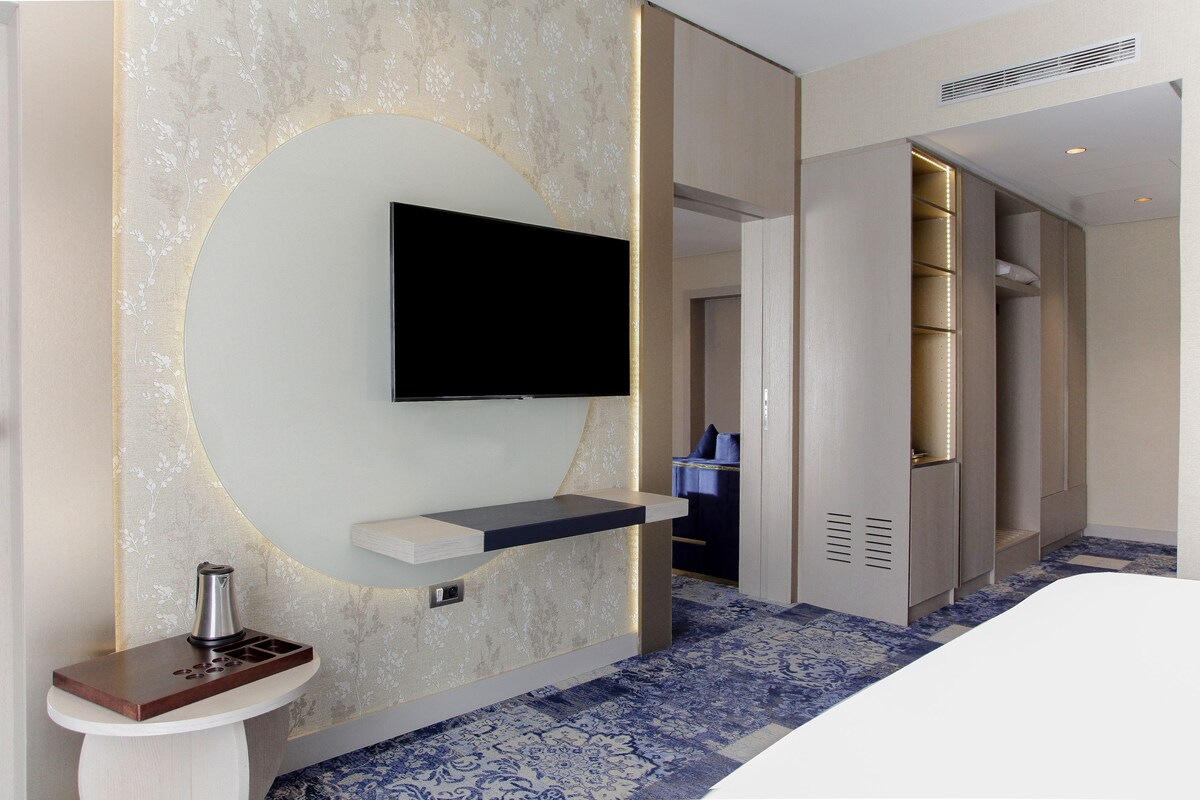 A modern hotel room with a flat-screen TV mounted on the wall, a small bedside table, and a walk-in closet.