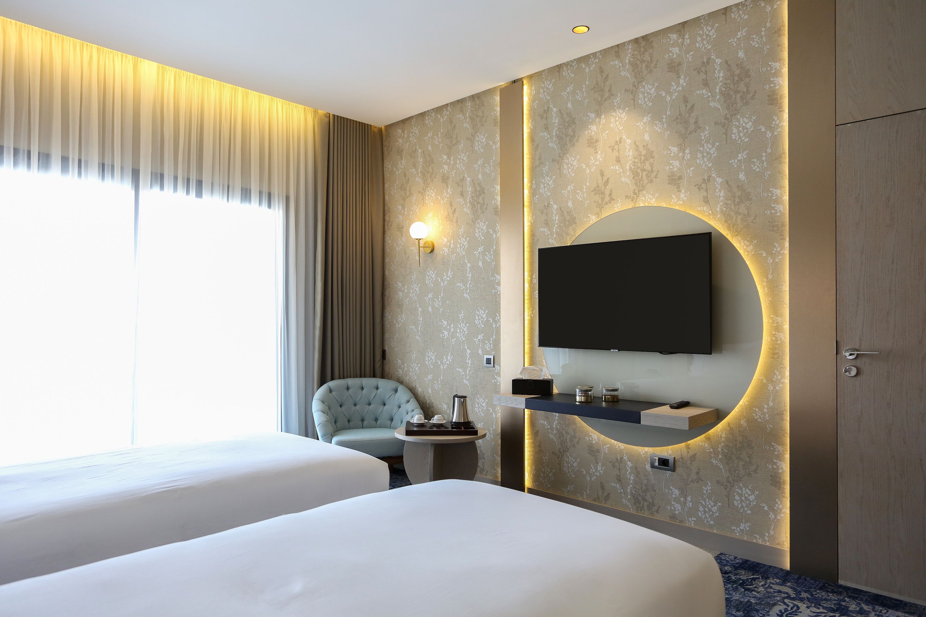Habitación de hotel con dos camas, un televisor montado en la pared, una mesita con una cafetera, una silla y un amplio ventanal con cortinas.