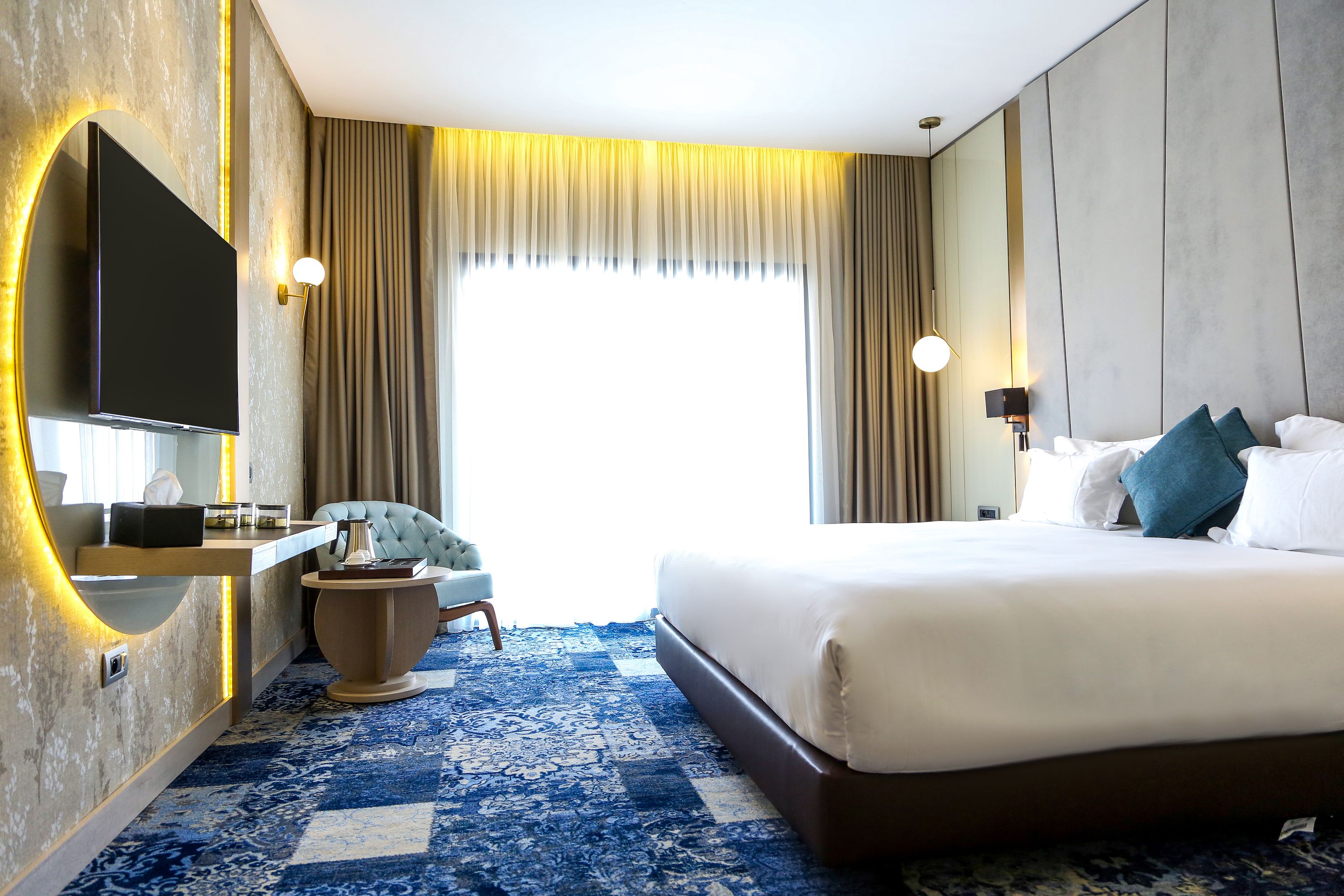 Ein Hotelzimmer mit einem großen Bett, einem an der Wand befestigten Fernseher, einem Schreibtisch mit Stuhl und einem großen Fenster mit Vorhängen.