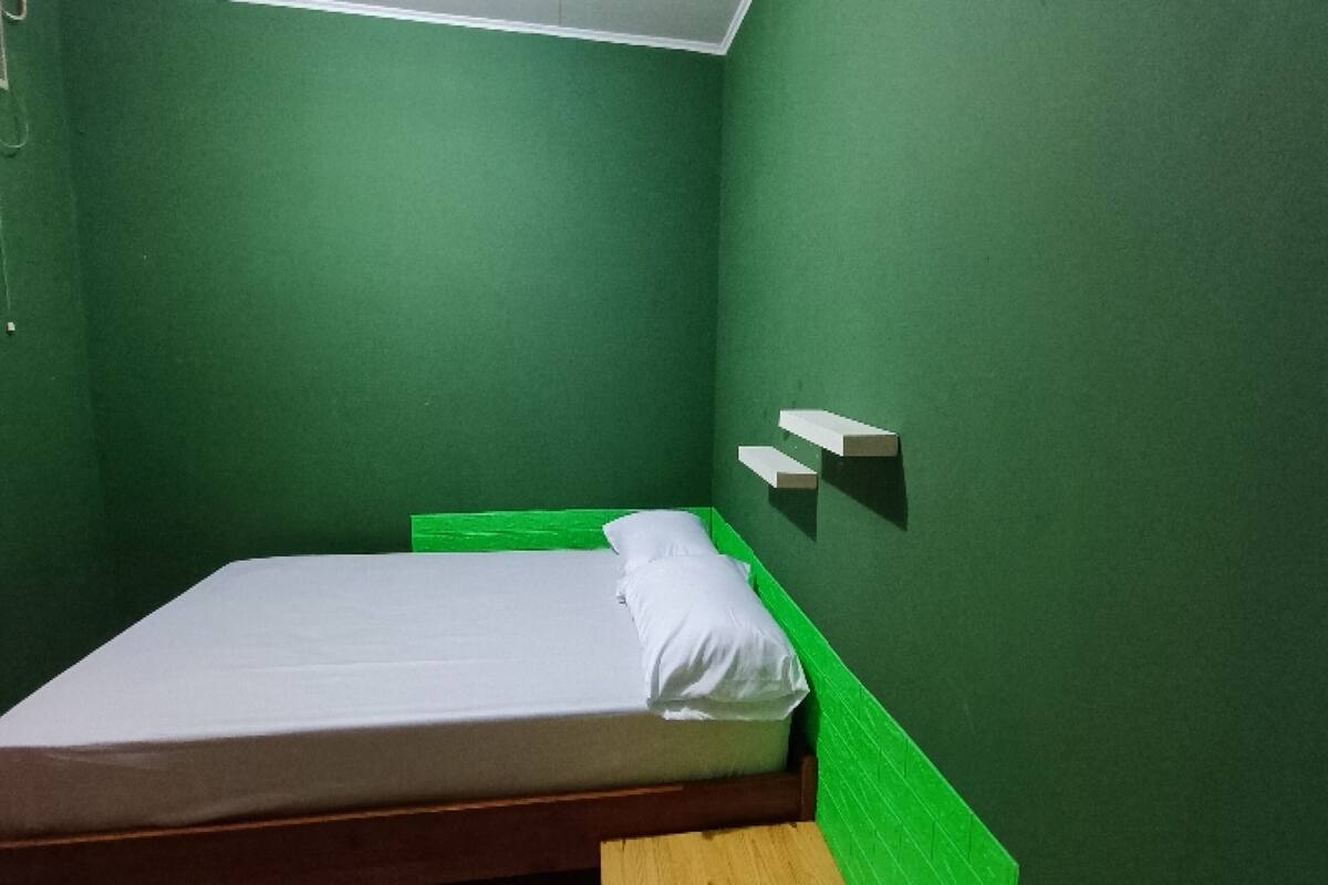 Una habitación pequeña con cama individual, paredes verdes, cabecera de madera y colchón blanco.