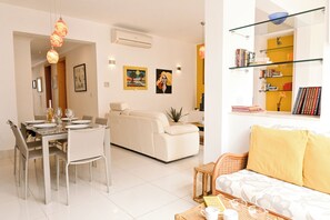 A modern living room with a sofa, dining table, and chairs, a bookshelf, and a small side table with a plant. - Modern & Terrace by the beach, 3brd in Qawra, Qawra - Saint Paul's Bay, Malta (Qawra - Saint Paul's Bay)