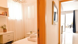 A hotel room with a bed, a bedside table with a plant, a small table with a lamp, and a view of the outside. - Modern & Terrace by the beach, 3brd in Qawra, Qawra - Saint Paul's Bay, Malta (Qawra - Saint Paul's Bay)