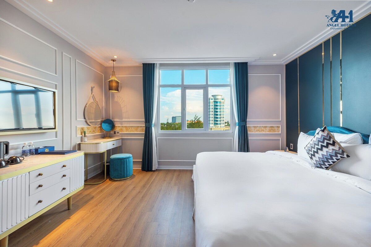 Ein Hotelzimmer mit Bett, Schreibtisch, Schminktisch, Spiegel, einem Fenster mit Vorhängen und Blick auf Gebäude.