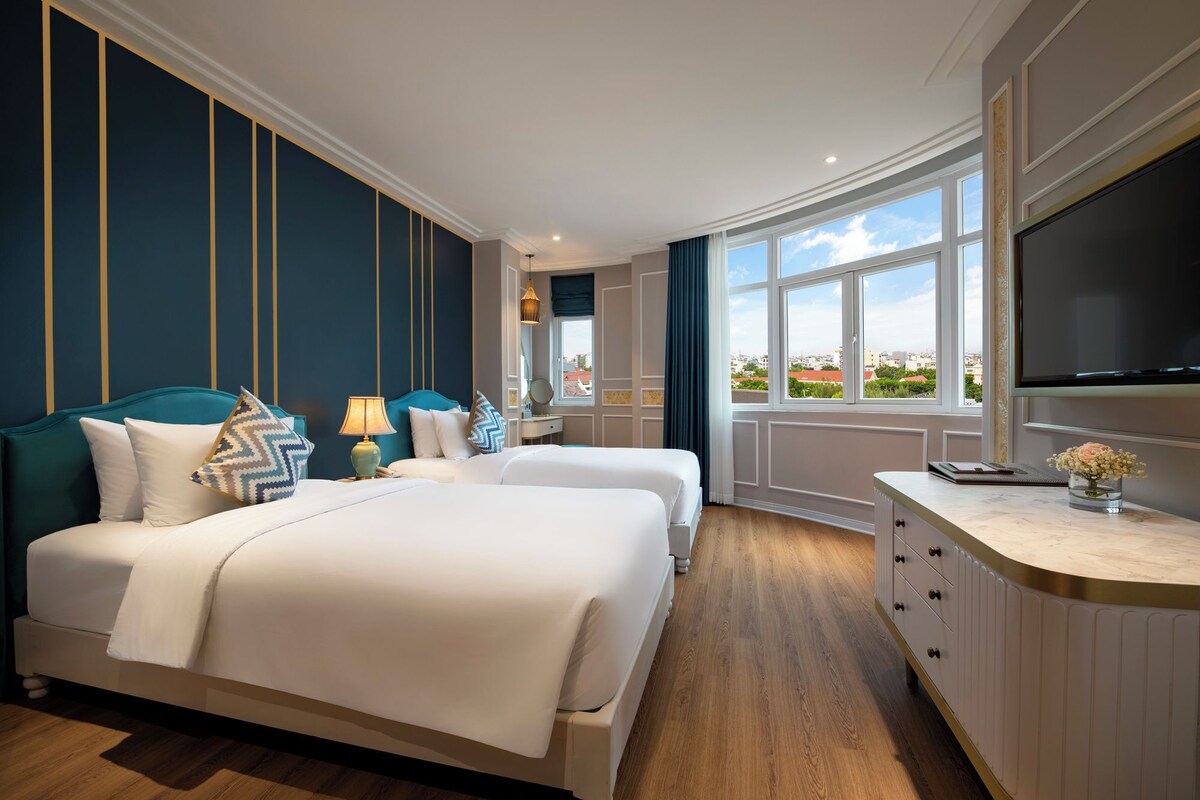 Ein Hotelzimmer mit zwei Betten, einem Fernseher, einem Schreibtisch und einem Fenster mit Blick auf die Stadt.