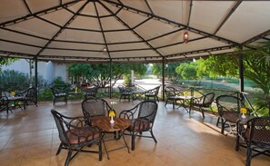 An outdoor patio with tables and chairs, surrounded by greenery and a view of a park. - Club Mahindra Gir (Mendarda)