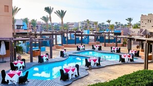 A poolside area with multiple dining tables and chairs, surrounded by a pool and palm trees.