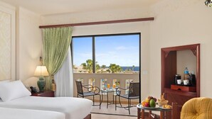 A hotel room with a balcony, a bed, a dining table with chairs, and a small table with fruits and a bottle.