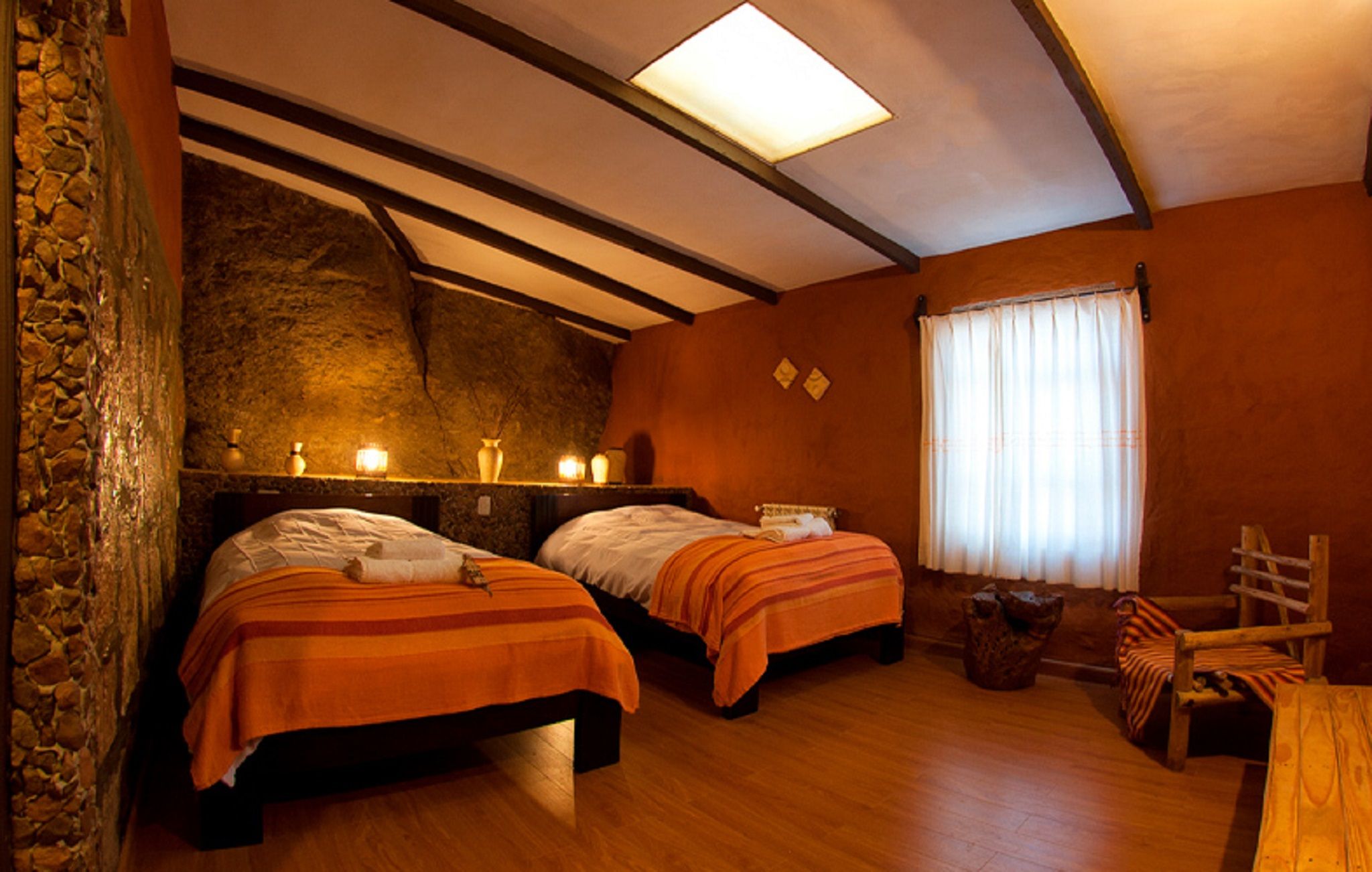 Una habitación con dos camas, pared de piedra, techo de madera y una ventana con cortinas.