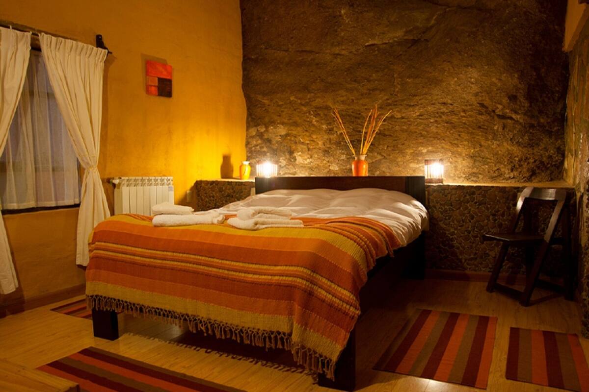 Un dormitorio con pared de piedra, cama con manta a rayas, suelo de madera y ventana con cortinas.