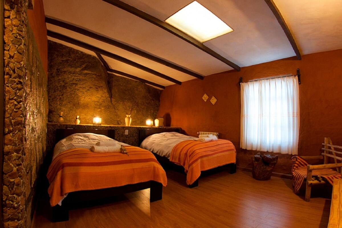 Una habitación con dos camas, pared de piedra, techo de madera y una ventana con cortinas.