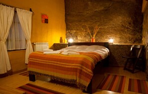 A bedroom with a stone wall, a bed with a striped blanket, a wooden floor, and a window with curtains.