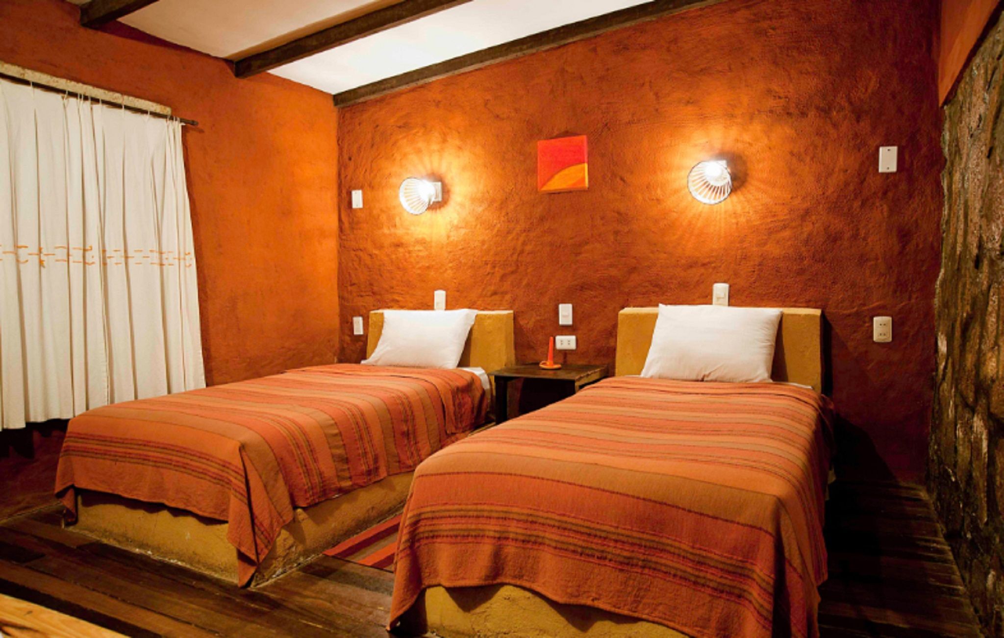 Una habitación con dos camas, suelo de madera, un ventanal con cortinas y un aplique en la pared.