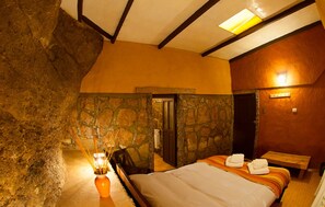 A bedroom with a stone wall, a bed with towels, a wooden bench, and a small table with a candle.
