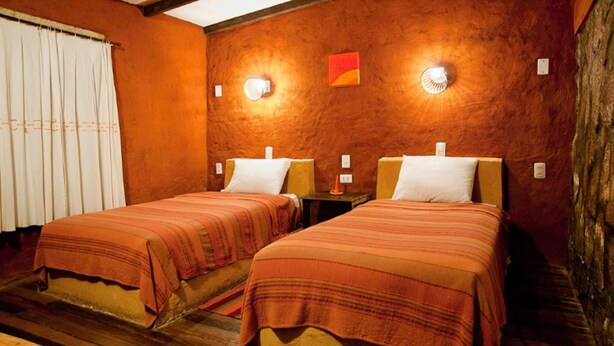 A room with two beds, wooden flooring, a window with curtains, and a wall-mounted light fixture. - Hotel Jardines de Mallku Cueva (Colcha K)