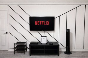 A modern living room with a flat-screen TV mounted on a wall, a black entertainment unit below, and a standing heater to the right. - Clean & Cozy 2BR Raleigh Stay Book Your Dates Now (Raleigh)