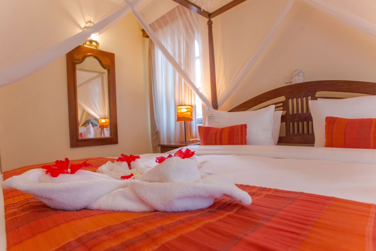 A four-poster canopy bed with white linens and red accents, a mirror, and a nightstand with a lamp.