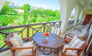 A balcony with a wooden table, chairs, and a view of a lush garden. - Mzima Beach Resort (Diani Beach)