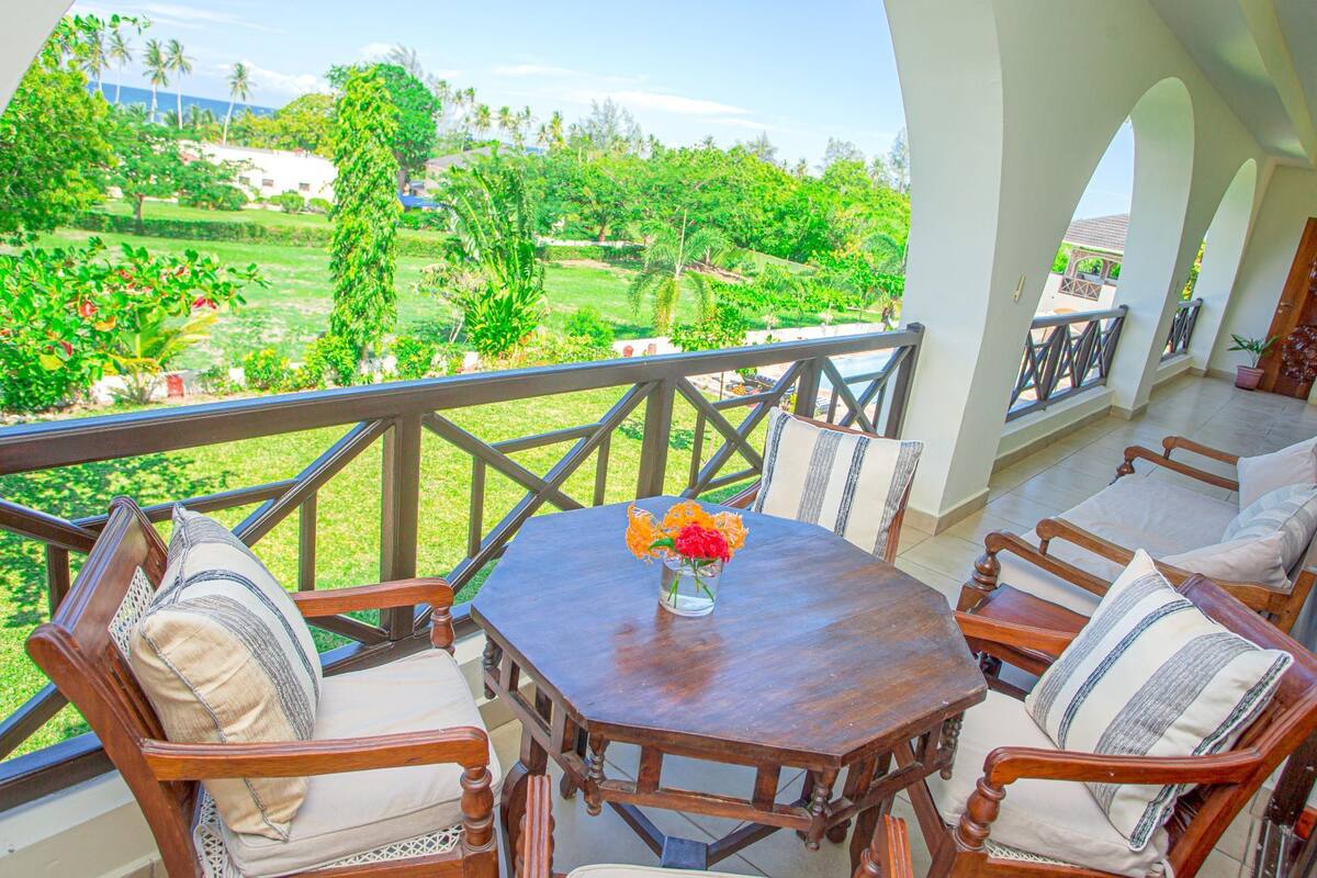 A balcony with a wooden table, chairs, and a view of a lush garden.