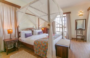 A four-poster bed with a canopy, a bedside table, a nightstand, a bedside lamp, a bedside mirror, and a window with curtains. - Mzima Beach Resort (Diani Beach)