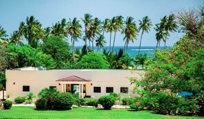 A building with a flat roof, surrounded by lush greenery and palm trees, overlooking a body of water. - Mzima Beach Resort (Diani Beach)