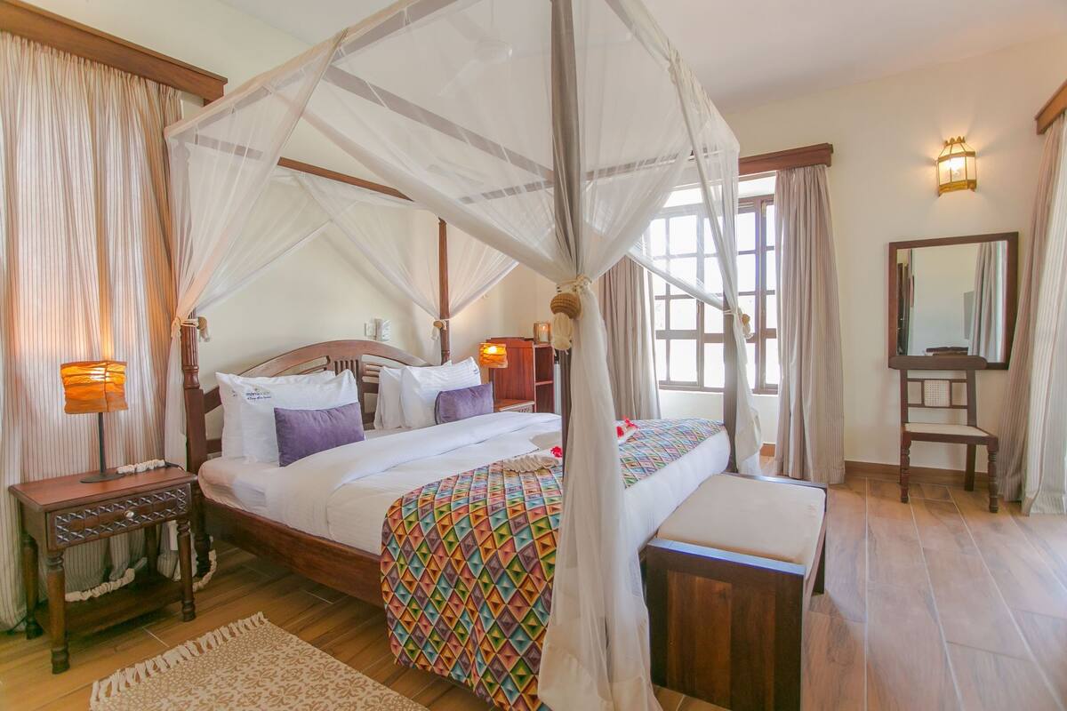 A four-poster bed with a canopy, a bedside table, a nightstand, a bedside lamp, a bedside mirror, and a window with curtains.