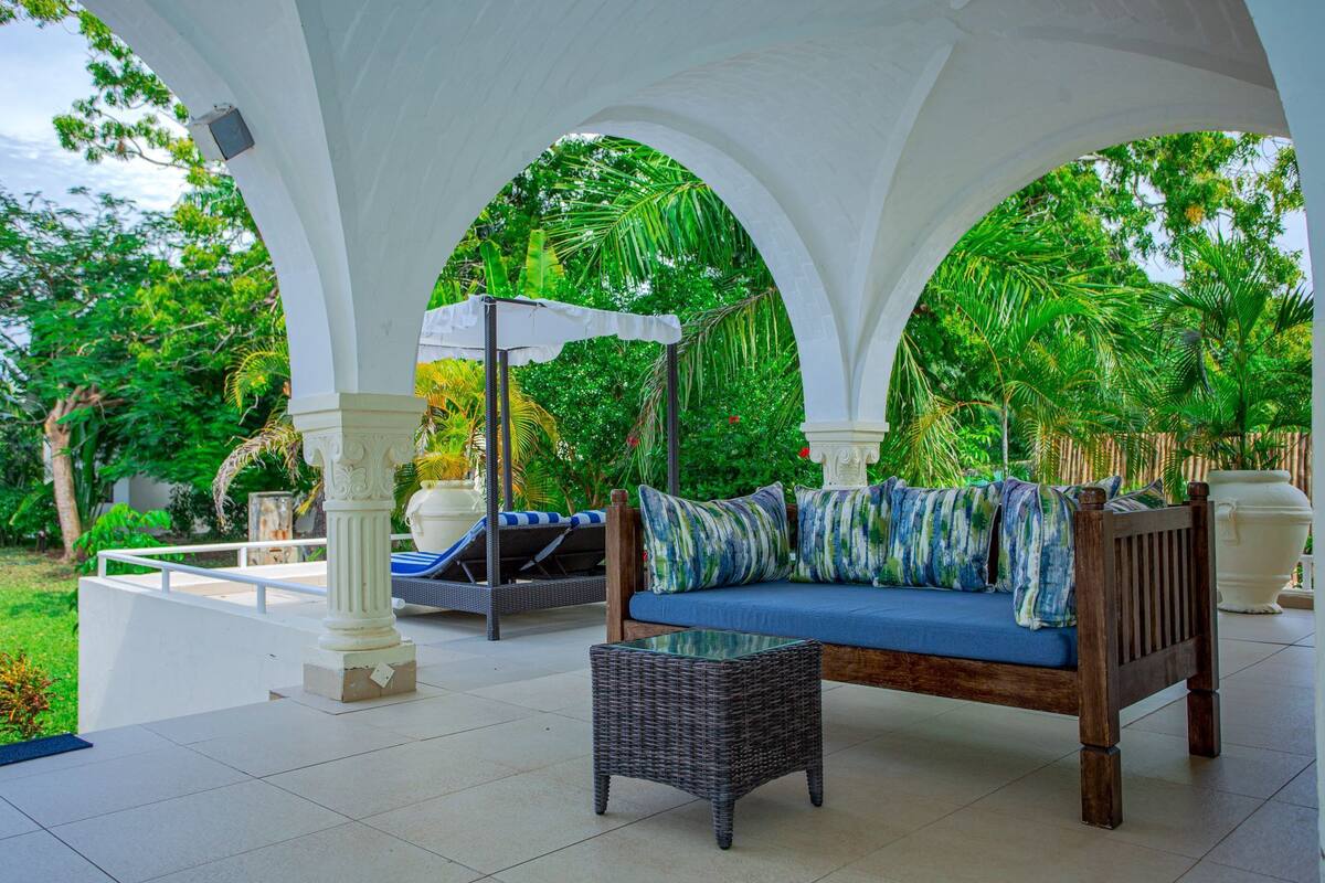 A covered outdoor seating area with a sofa and a table, surrounded by white columns and lush greenery.