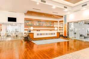 A hotel reception area with a wooden reception desk, a large colorful mural, and a glass door entrance. - Club Mahindra Jaipur (Jalsu)