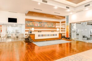 A hotel reception area with a wooden reception desk, a large colorful mural, and a glass door entrance.
