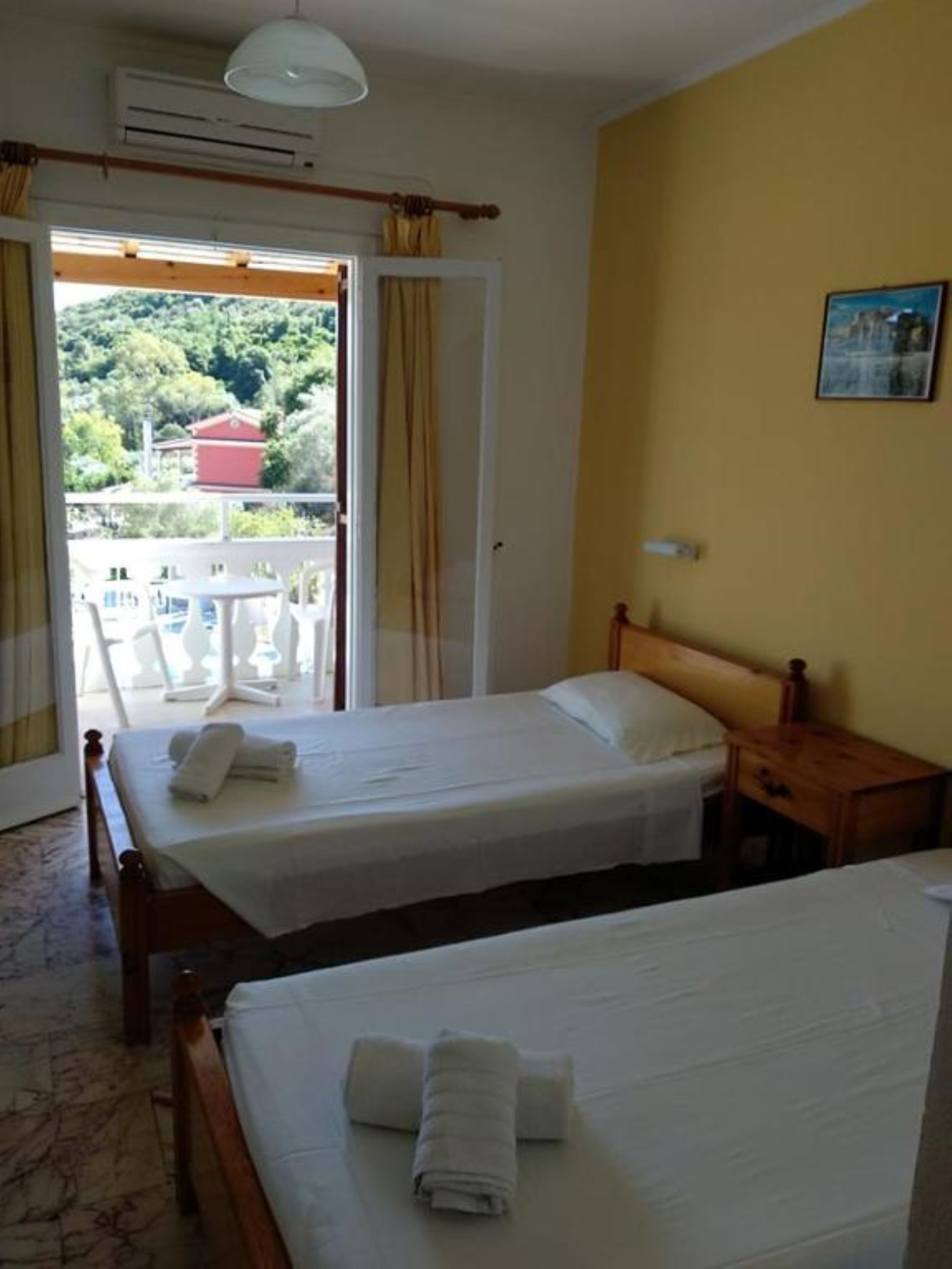 A hotel room with two beds, a wooden headboard, a small bedside table, and a view of greenery through an open door.