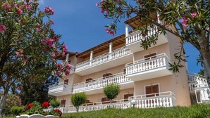 A multi-story building with balconies, a garden with pink flowers, and a clear blue sky. - Corfu Panorama (Corfu)