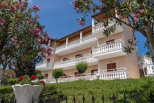 A multi-story building with balconies, a garden with pink flowers, and a clear blue sky.