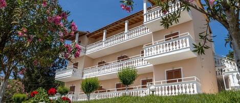 Un edificio de varios pisos con balcones, un jardín con flores rosadas y un cielo azul despejado.