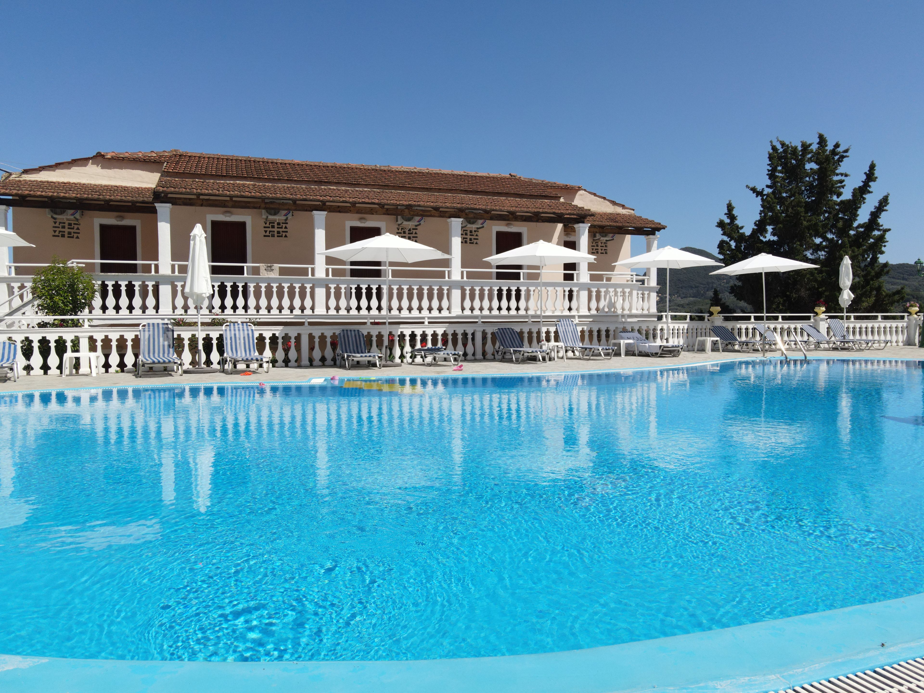 A swimming pool with lounge chairs and umbrellas, a building with a tiled roof, and a white fence.
