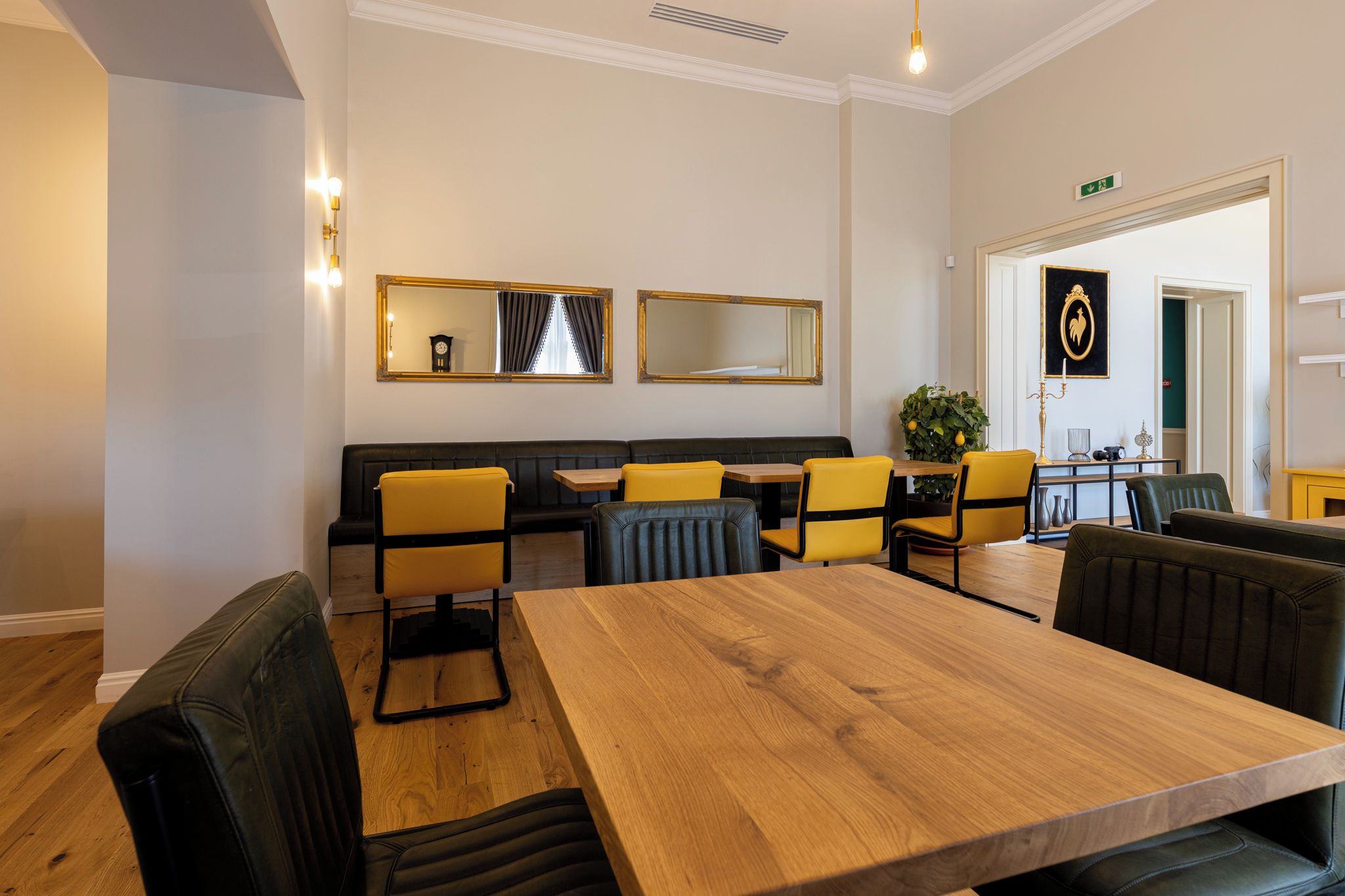 A hotel room with a wooden dining table, yellow chairs, a black sofa, and a mirror on the wall.