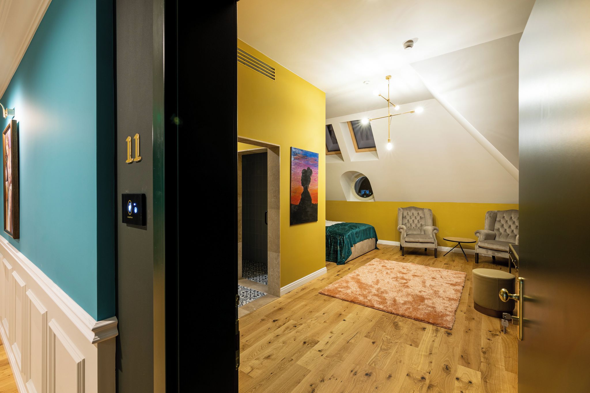 Quarto com piso de madeira, parede verde-água, parede amarela, um quadro, luminária no teto e uma porta com número.