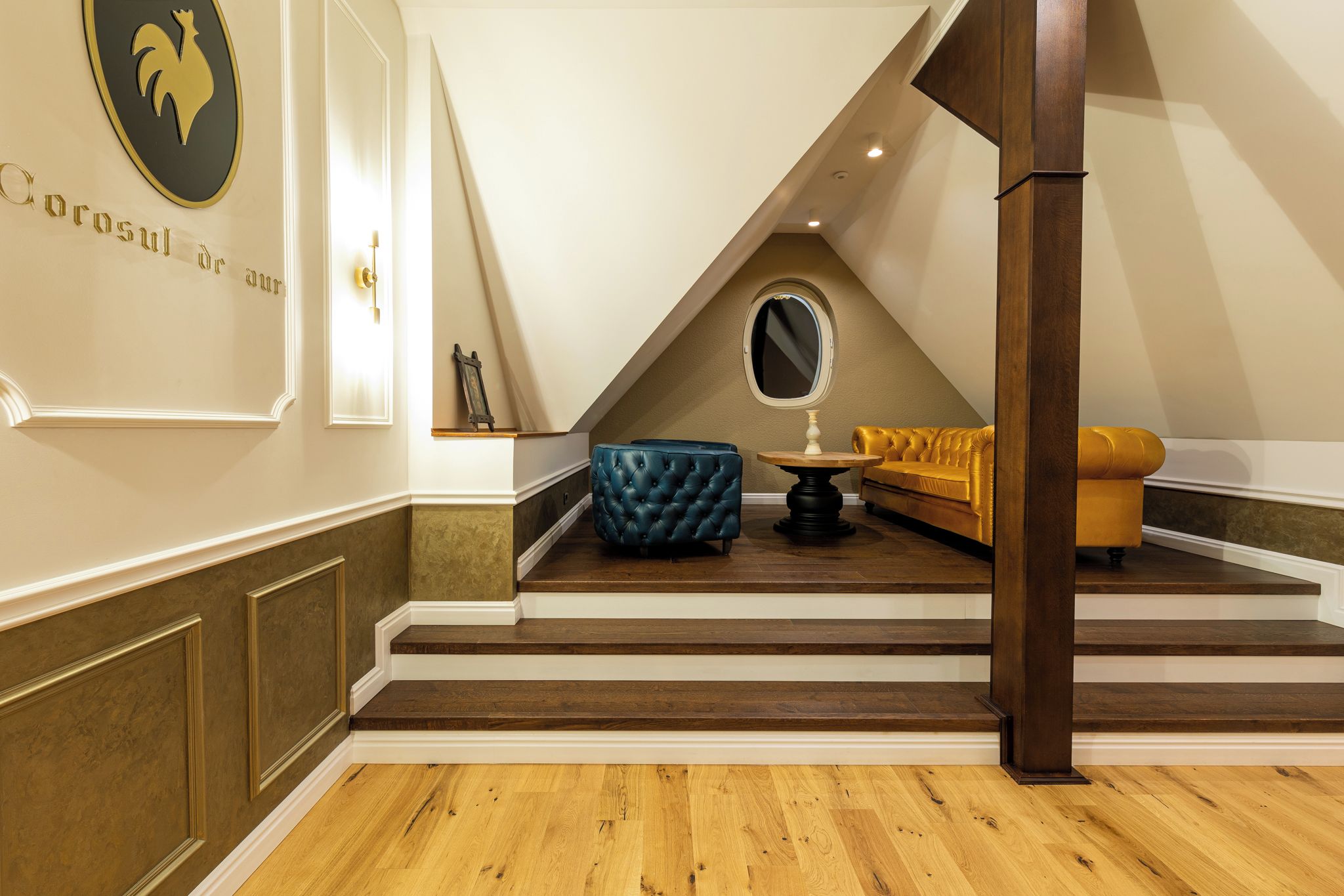 Ein Zimmer mit Holzboden, einem blauen, mit Wirbeln versehenen Ottomanen, einer goldenen Couch, einem runden Spiegel und einem an der Wand befestigten Wappen.