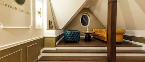 A room with a wooden floor, a blue tufted ottoman, a gold sofa, a round mirror, and a wall-mounted emblem.