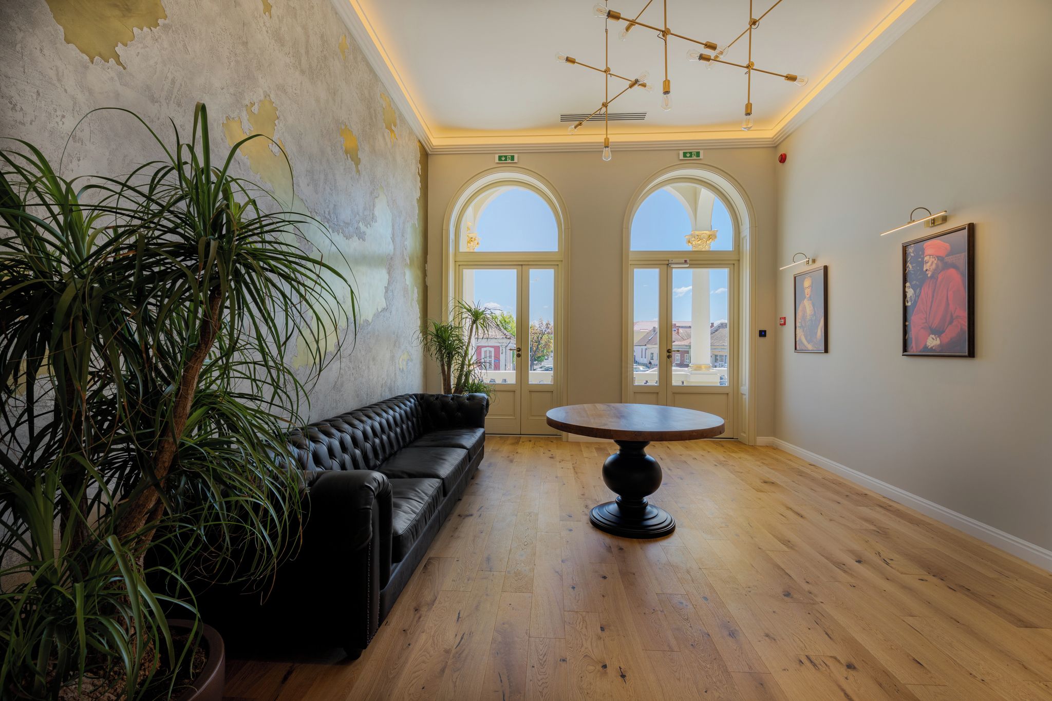 A room with a black leather sofa, a round table, and two arched windows with views outside.